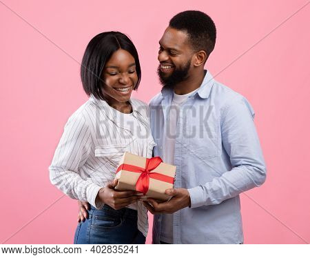 Young Married Couple Celebrating Valentines Day, Holding Wrapped Gift Box On Pink Studio Background.