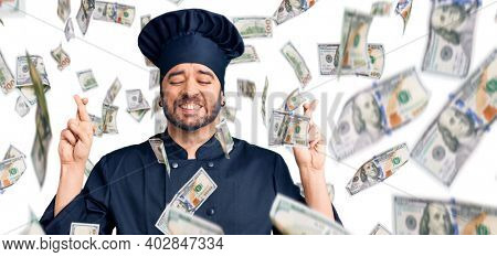 Young hispanic man wearing cooker uniform gesturing finger crossed smiling with hope and eyes closed. luck and superstitious concept.