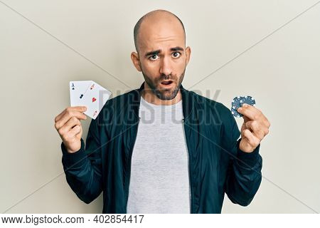 Young hispanic man playing poker holding casino cards and chips in shock face, looking skeptical and sarcastic, surprised with open mouth 