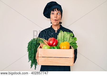Beautiful brunettte woman chef holding fresh veggetables making fish face with mouth and squinting eyes, crazy and comical. 