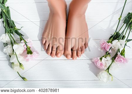 Top View Of Female Feet With Pastel Pink Toenails Near Carnation And Eustoma Flowers On White Wooden