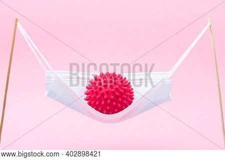 A Red Ball With Spikes Coronavirus Swinging In A Medical Mask Like In A Hammock Or Swing.