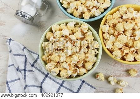 High angle shot of three different colored bowls of fresh popped popcorn with towl and salt shaker with salt spilling out.