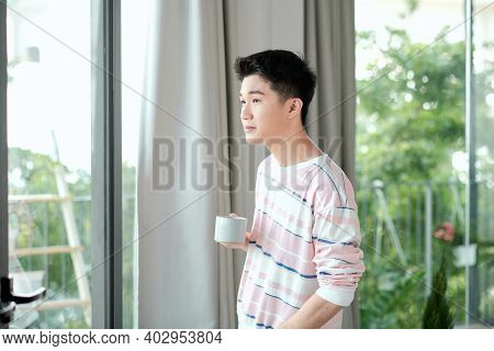 Young handsome carefree man near modern full length window enjoying a cup of coffee while looking outside