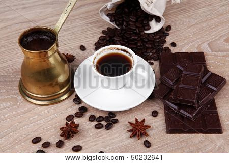 sweet hot drink : black Turkish coffee in small white mug with coffee beans spilled on a wooden table with stripes of dark chocolate and copper Arab Cezve full coffee
