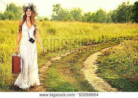 Romantic smiling young woman in a circlet of flowers goes on a country road with her old camera and suitcase.