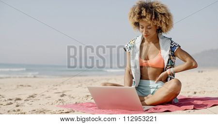 African-american woman sits on a towel in the sand with laptop on the summer beach