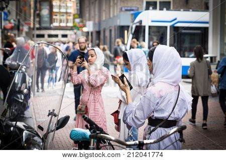AMSTERDAM HOLLAND - AUGUST 21 2017; Three young Muslim women in white hijab in busy city street standing pre-occupied using mobile phones