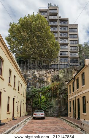 Sydney Australia - March 22 2017: The cul-de-sac short Adherden street end against a cliff. Yellow painted houses. On top of cliff modern high rise and big green tree. Can in street.