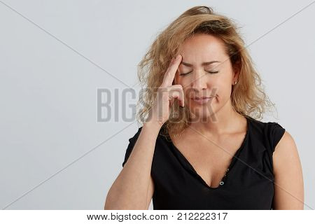 Emotional beautiful young adult woman portrait. Studio shoot. Isolated on White Background. A woman in a black T-shirt with a deep neckline holds two fingers at her temple. She has a headache.