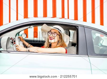 Poortrait of a young happy woman driving a car on the red wall background during the summer vacation