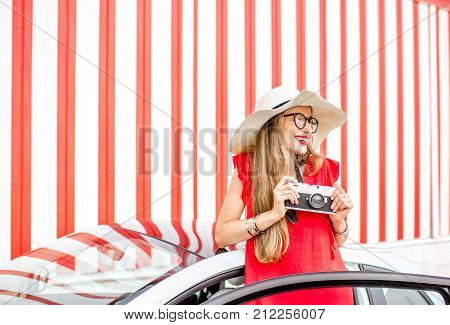 Poortrait of a young happy woman pulling out of the car door with photo camera on the red wall background during the summer vacation