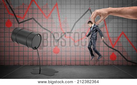 A small businessman caught by a giant male hand over a spilling oil barrel and red negative economic graphs. Saved from crisis. Oil and gas business. Prices fallen down.