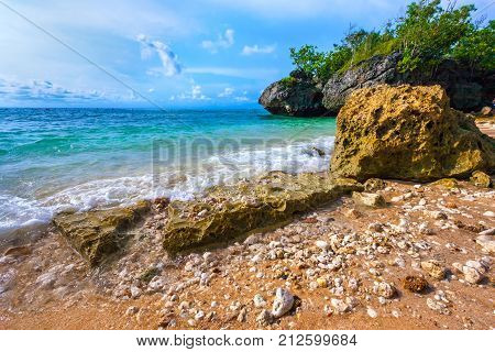 Close-up of stones with a wave on the beach against a rock background and a sunny blue sky with white clouds. Padang Padang Beach, Bali, Indonesia.