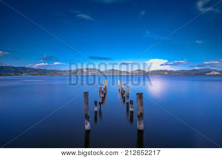Wooden pier or jetty remains on blue lake sunset and sky reflection water. Long exposure Versilia Massaciuccoli Tuscany Italy.