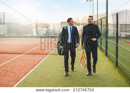 Business, partnership and sport. Front view successful businessman walking and chatting after a tennis set, wearing suits and tennis equipment.  Business partners practicing sport together. Mentoring.