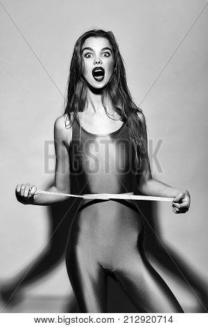 Attractive slim girl with surprised face and long hair in violet second skin jumpsuit standing with tape-line around waist on yellow background