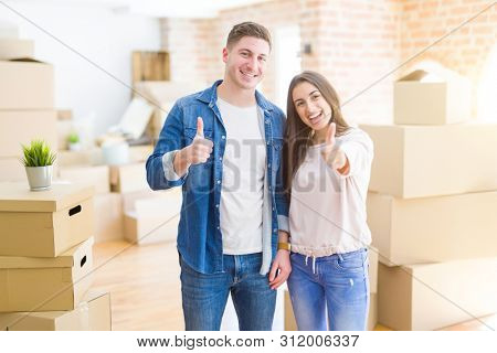 Beautiful young couple moving to a new house doing happy thumbs up gesture with hand. Approving expression looking at the camera with showing success.
