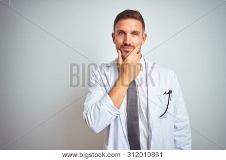 Young handsome doctor man wearing white profressional coat over isolated background looking confident at the camera with smile with crossed arms and hand raised on chin. Thinking positive.