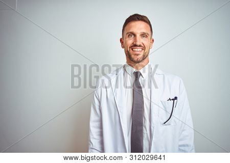 Young handsome doctor man wearing white profressional coat over isolated background with a happy and cool smile on face. Lucky person.