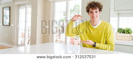 Wide angle shot of young handsome man at home gesturing with hands showing big and large size sign, measure symbol. Smiling looking at the camera. Measuring concept.
