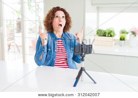 Senior woman doing video chat using smartphone camera amazed and surprised looking up and pointing with fingers and raised arms.