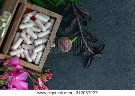Close Up Image Of Herbal Capsules In Old Wooden Box With Pink Flower, Wild Fruit And Herbs On Dark B