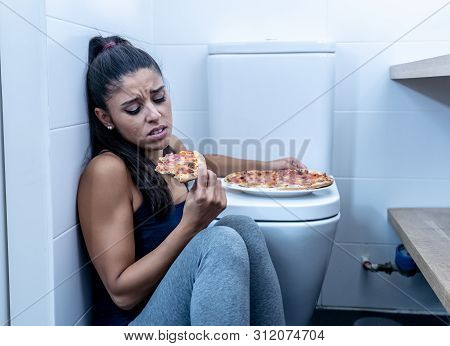 Young Beautiful Bulimic Woman Sitting On The Bathroom Floor Eating Pizza Looking Guilty