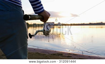 A Man In Striped Shirts With Fishing Rods Staying On The Riverside At Sunset