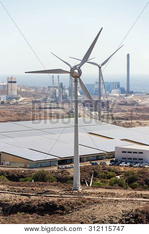 White Turbines At Wind Farm With Power Station And Sea In Background, Arid Landscape On Sunny Summer