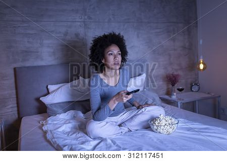 Beautiful Young Mixed Race Woman Wearing Pajamas Sitting On Bed, Eating Popcorn And Changing Channel