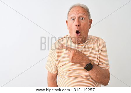 Senior grey-haired man wearing striped t-shirt standing over isolated white background Surprised pointing with finger to the side, open mouth amazed expression.
