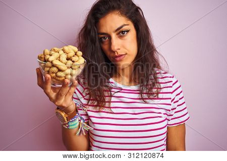 Young beautiful woman holding bowl with peanuts over isolated pink background with a confident expression on smart face thinking serious