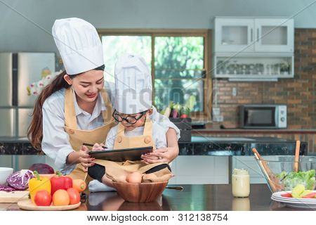 Young Woman And Kid Using Tablet Computer While Cooking In Kitchen. Householding, Tasty Food And Dig