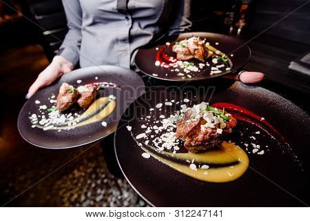Woman Waiter Carries Three Plates Of Food. Meat Steak Garnished With Two Sauces On A Dark Plate. Clo