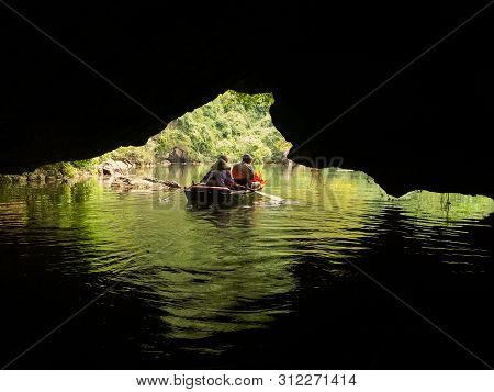 Boat Filled With Tourists And Local Guide In Bamboo Hat Exploring Caves On A River Surrounded By Bea