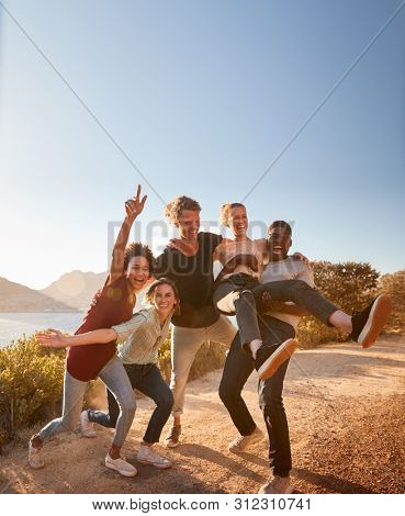 Five millennial friends on a road trip have fun posing for photos on a coastal path, full length