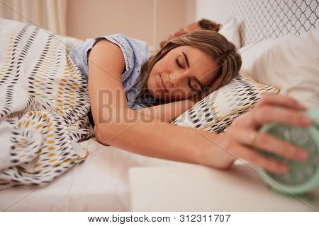 White woman half asleep in bed reaches out for alarm clock, partner sleeping in background, close up