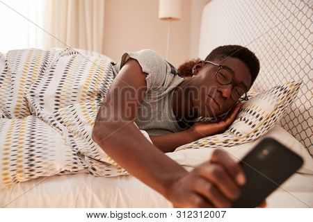 Millennial African American man wearing glasses, half asleep in bed holding smartphone, close up
