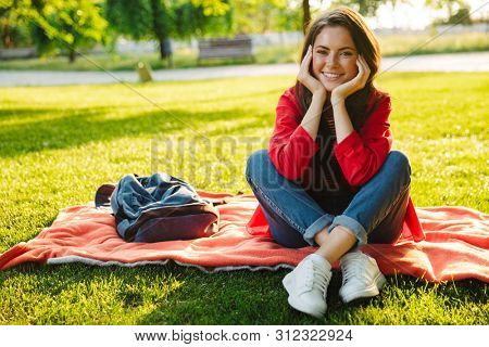 Image of nice girl student wearing red jacket smiling and propping up her head while sitting on blanket in green park