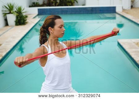 Front view of fit mixed-race woman exercising with resistance band near swimming pool in the backyard. Summer fun at home by the swimming pool