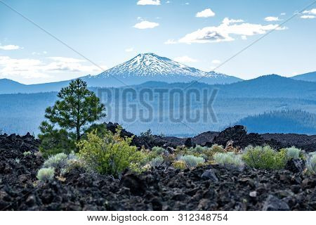 View Of Mt. Bachelor From Newberry Volcano National Monument. Black Lava Rocks, Trees And Desert Veg