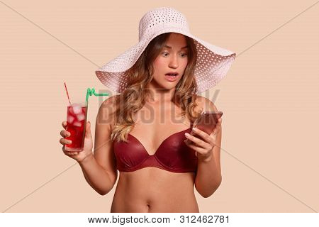 Shocked Emotional Model Posing With Wide Opened Eyes And Mouth, Holding Smartphone And Cold Drink In