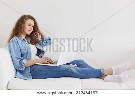 Horizontal Shot Of Good Looking Curly Haired Young Model Sitting On White Sofa, Having Laptop, Watch