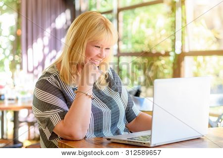 Happy Middle Age Woman Sitting In A Coffee Shop And Using A Laptop Computer, Smiling, Chatting Onlin