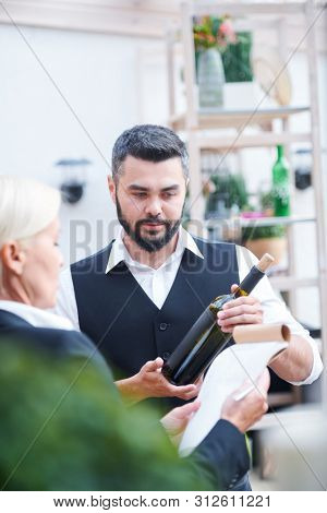Young bartender giving characteristics of new sort of wine in bottle to cavist