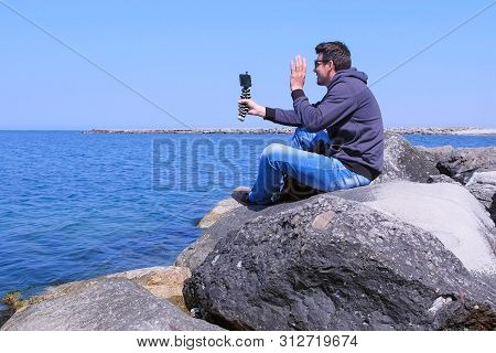 Man Traveller Talks Video Chat On Smartphone Sits On Sea Stone Beach On Vacation. Mobile Phone With 