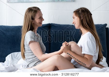 Side View Of Two Lesbians Holding Hands And Looking At Each Other While Sitting On Bed In Bedroom