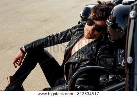 Handsome Young Man In Sunglasses Leaning On Motorcycle While Sitting On Ground