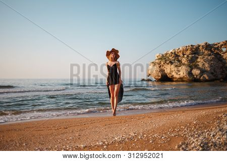 Beautiful Girl In A Black Dress And Hat Walks Along The Sea Sandy Shore At Sunset
